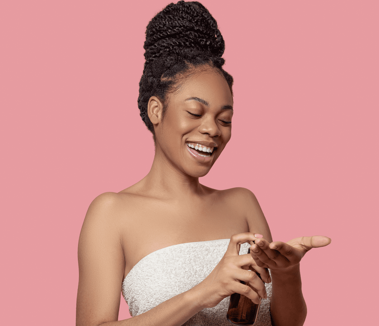 A woman in a towel applies lotion from a small bottle onto her hand, standing against a pink background.