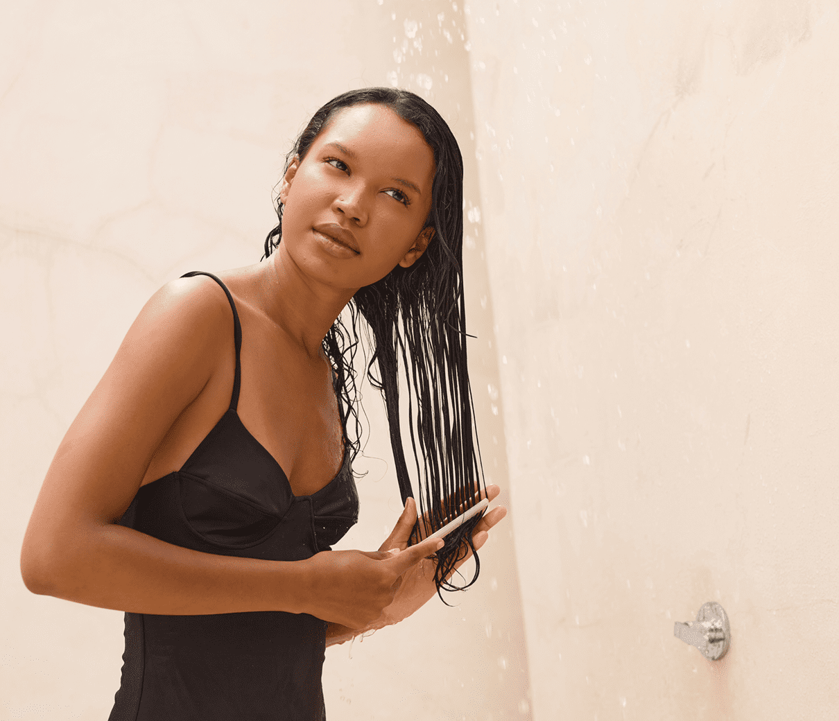 Person in a black swimsuit stands under a shower, brushing long wet hair with a contemplative expression.