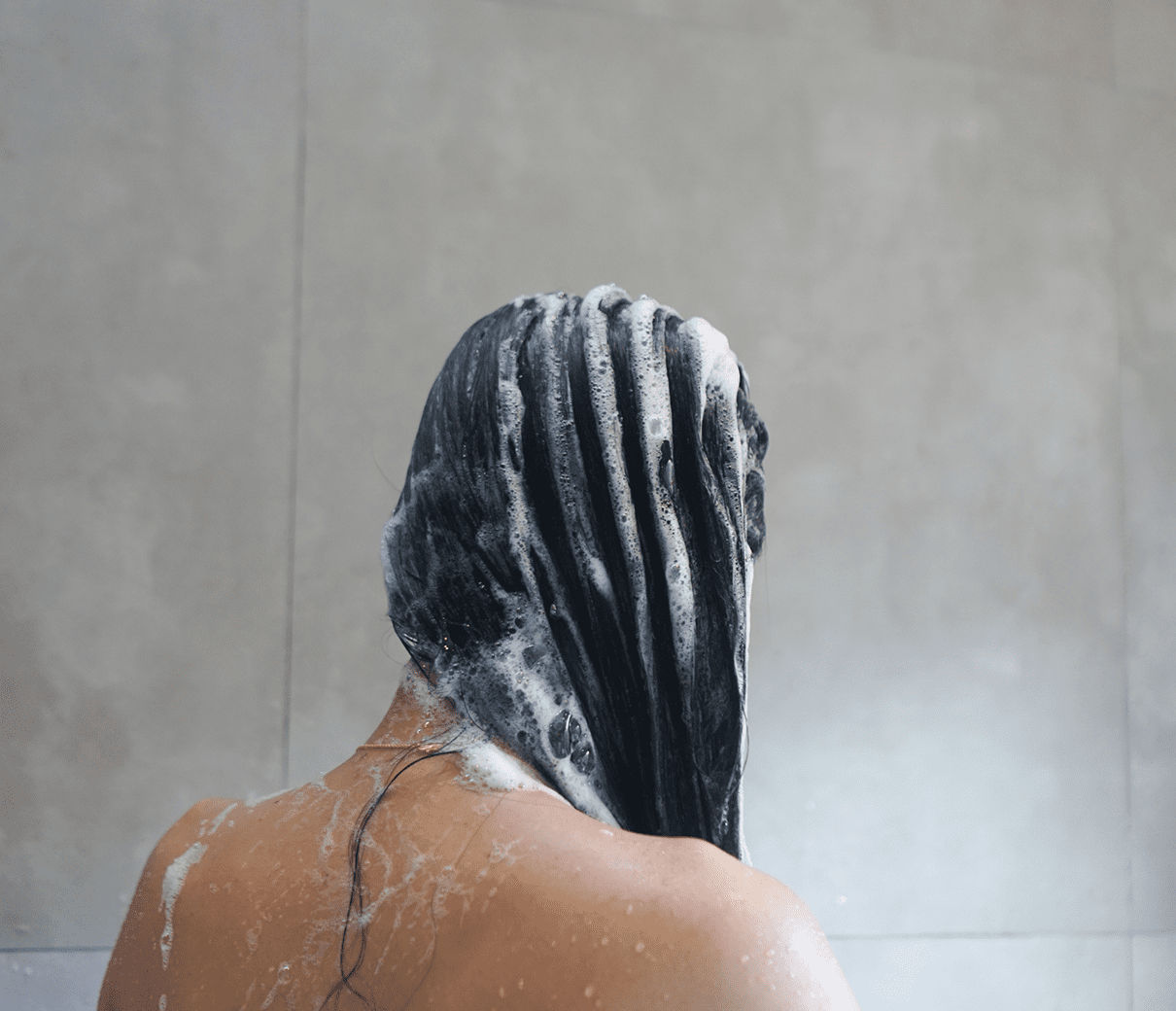 Person with long hair washing it, covered in suds, in a shower with a beige tiled wall.